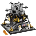 A lunar lander toy with two astronaut figures and a ramp is on a gray base with a flag.