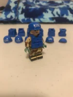 A toy figure wearing a blue helmet and vest with "UN" printed on them, with additional helmets and vests nearby.